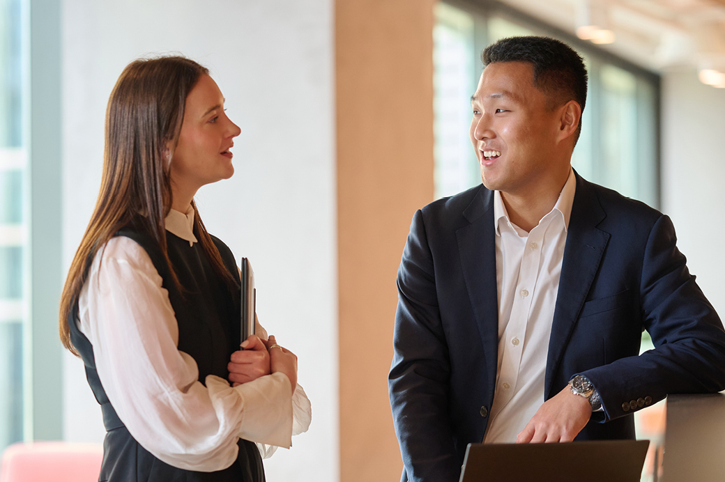Two employees stand together in a modern office, engaged in conversation beside a laptop. One person holds a laptop while listening to the other.