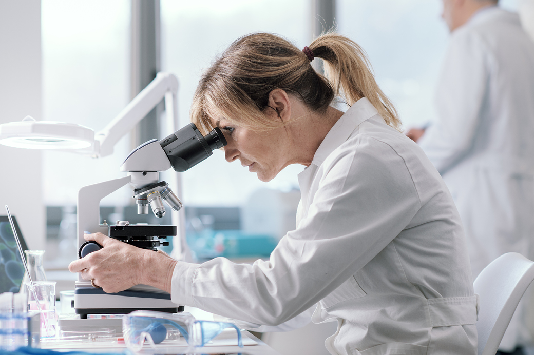 Female scientist with her hair in a ponytail wearing a white lab coat, carefully looking through a microscope in a bright laboratory.