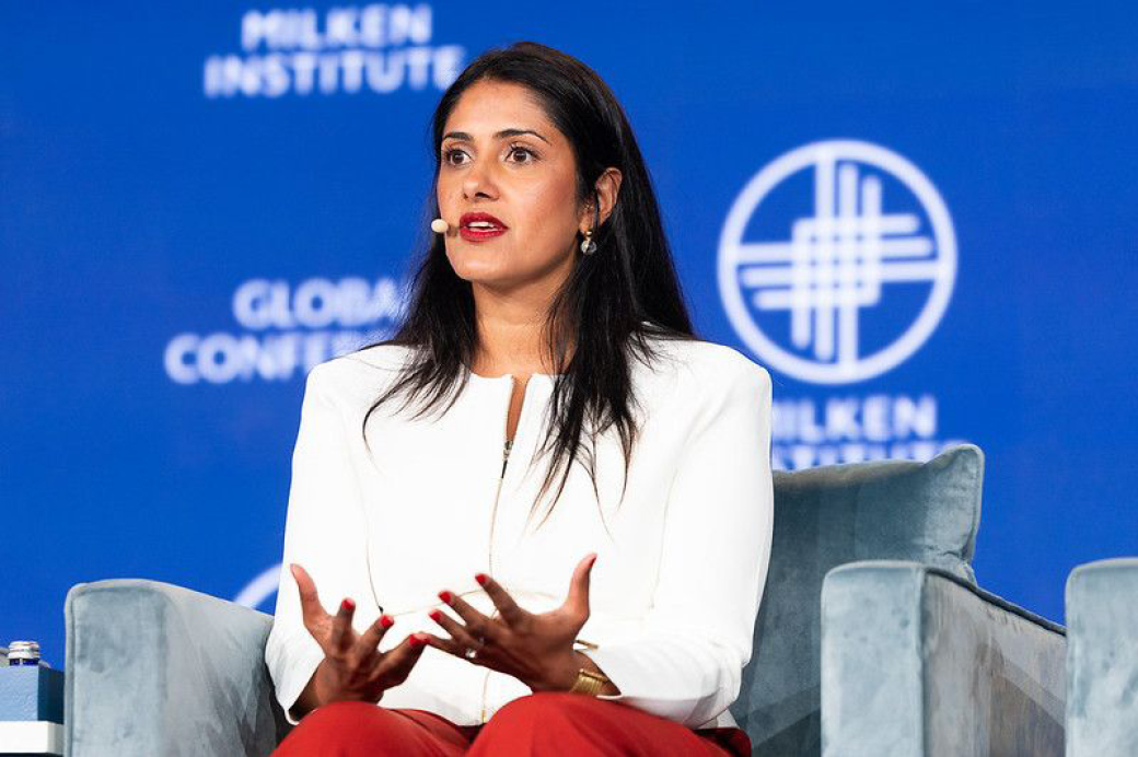 Shikha Gupta speaking while seated on a panel at a conference, in front of a Milken branded backdrop