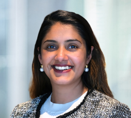 Professional headshot of Shikha smiling, wearing a patterned jacket and necklace, against a softly blurred office background.