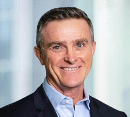 Professional headshot of Adam Tindall in a blue jacket and light blue shirt, photographed against a softly blurred office background.