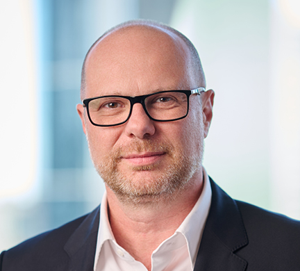 Professional headshot of Richard Brandweiner wearing glasses, a dark jacket and white shirt, photographed against a softly blurred office background.