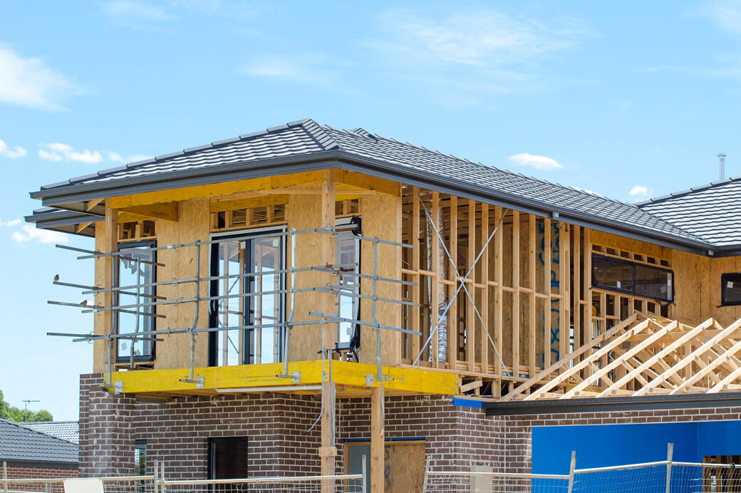 Residential homes under construction, showing timber framing, brickwork, scaffolding and roofing structures at a housing development site.