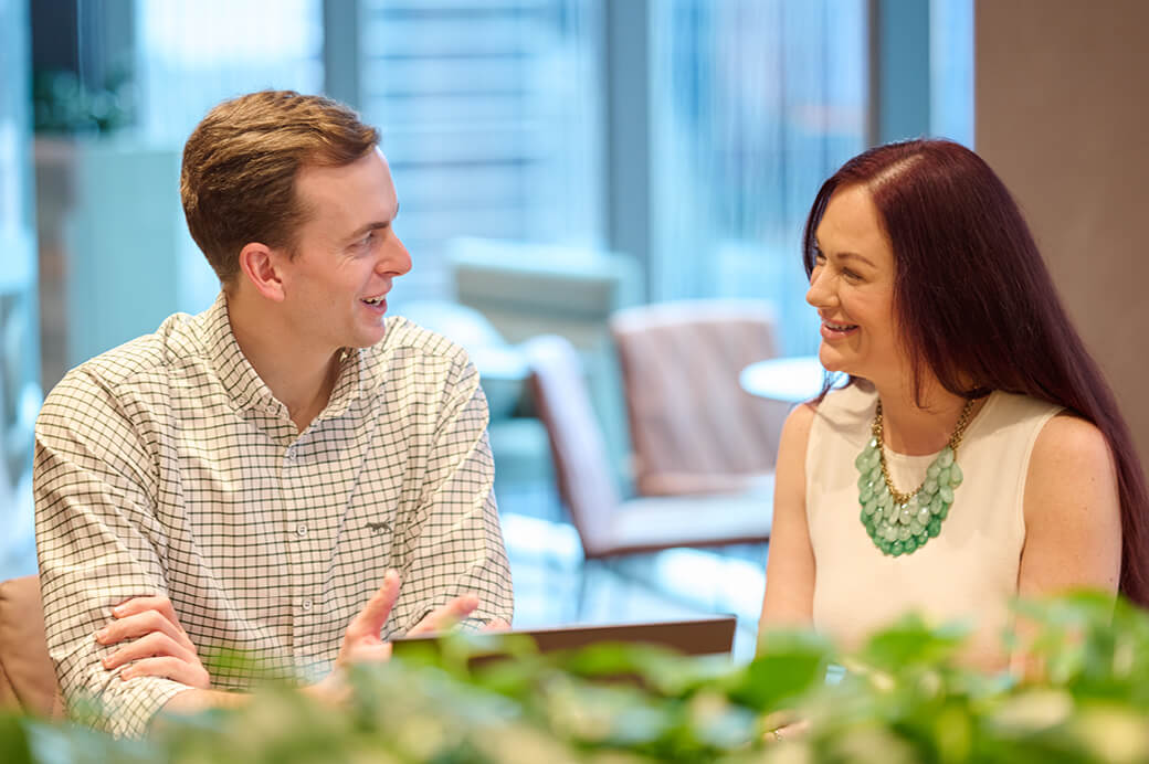 Two colleagues having a cheerful conversation in a modern office, with a man in a grid-check shirt speaking and a woman with reddish hair listening attentively.